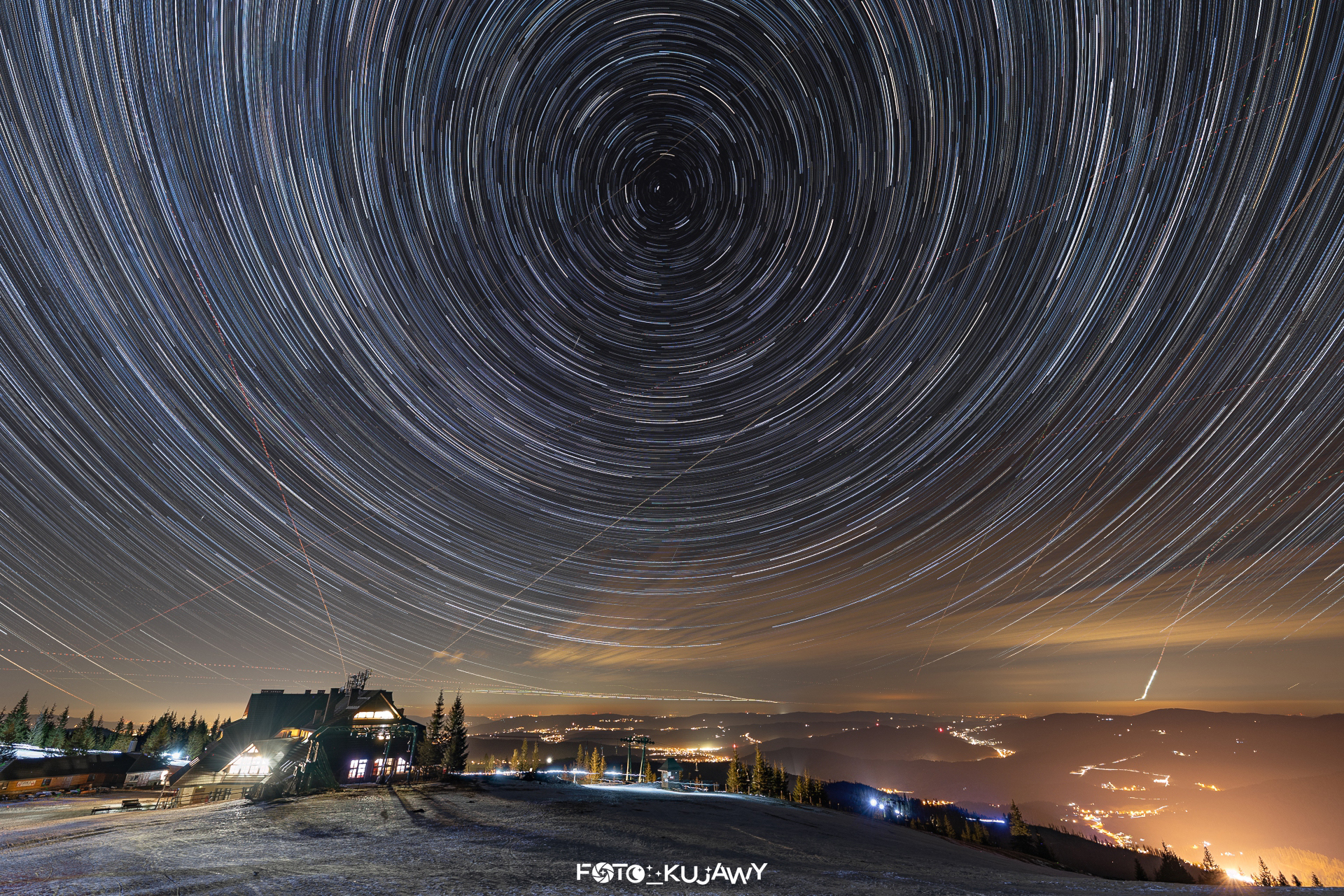 Niewysokie góry pokryte śniegiem w dolnej części zdjęcia, na górze gwiazdy ciągnące za sobą smugi i układające się w wir, którego centralny punkt znajduje się w środkowo-górnej części nieba. Smugi wytworzone przez gwiazdy gdzieniegdzie przecinają smugi wytworzone przez samoloty.