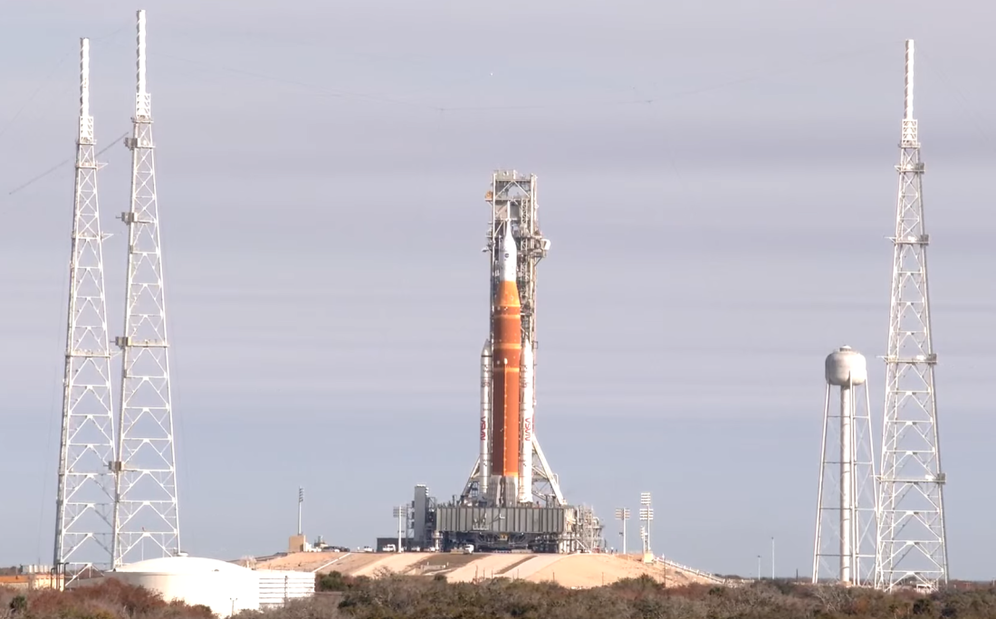 Wysoka rakieta Artemis II SLS z kapsułą Orion stoi na mobilnej platformie startowej na terenie kompleksu startowego 39B w Centrum Kosmicznym Kennedy’ego na Florydzie. Konstrukcję otaczają cztery wieże odgromowe, a rakieta przygotowywana jest do transportu z powrotem do budynku montażowego w celu przeprowadzenia prac technicznych.