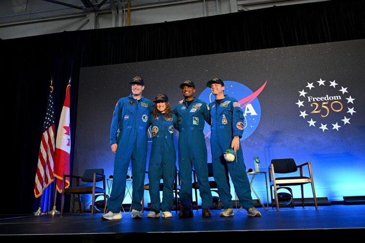 Cztery osoby w niebieskich kombinezonach NASA stoją na scenie z flagami USA i Kanady oraz logiem NASA w tle, obejmując się ramionami i uśmiechając do publiczności.