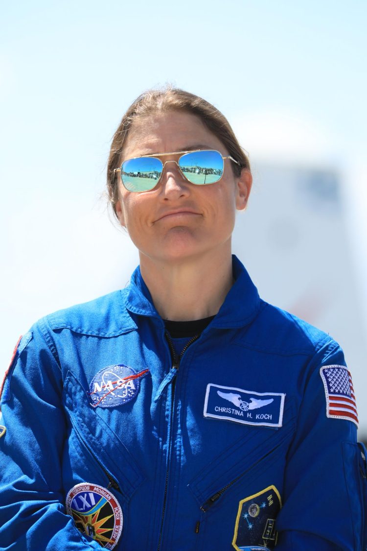 Stojąca na tle czystego nieba biała kobieta w okularach przeciwsłonecznych i niebieskim mundurze z logo NASA i innymi naszywkami.