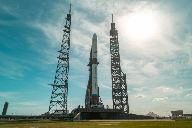 Biała rakieta pionowo stoi na stanowisku startowym, w tle niebieskie niebo delikatnie zachmurzone.