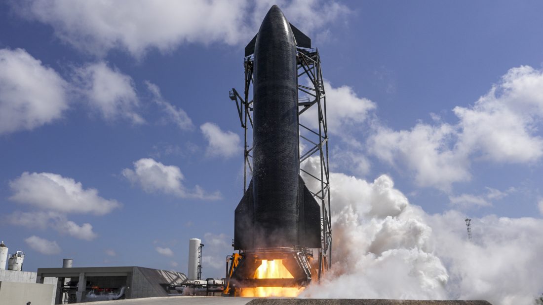 Widziana od spodu tylna część rakiety Starship z wlączonymi silnikami na tle niebieskiego nieba z kilkoma chmurami.