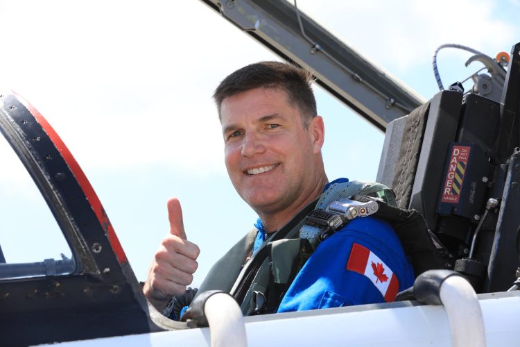 Biały mężczyzna siedzący w kokpicie myśliwca na tle czystego nieba, uśmiechający się do kamery, z naszywką flagi USA na ramieniu. Pokazuję gest kciuka w górę.