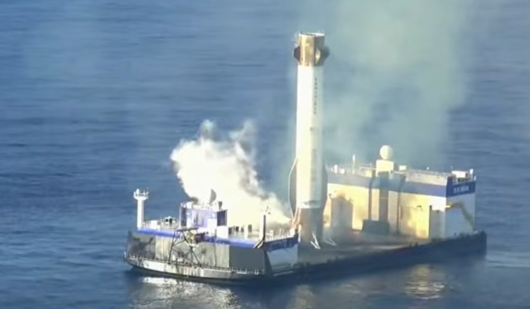 Biały pierwszy stopień rakiety stoi pionowo na pokładzie barki, dookoła spokojny ocean