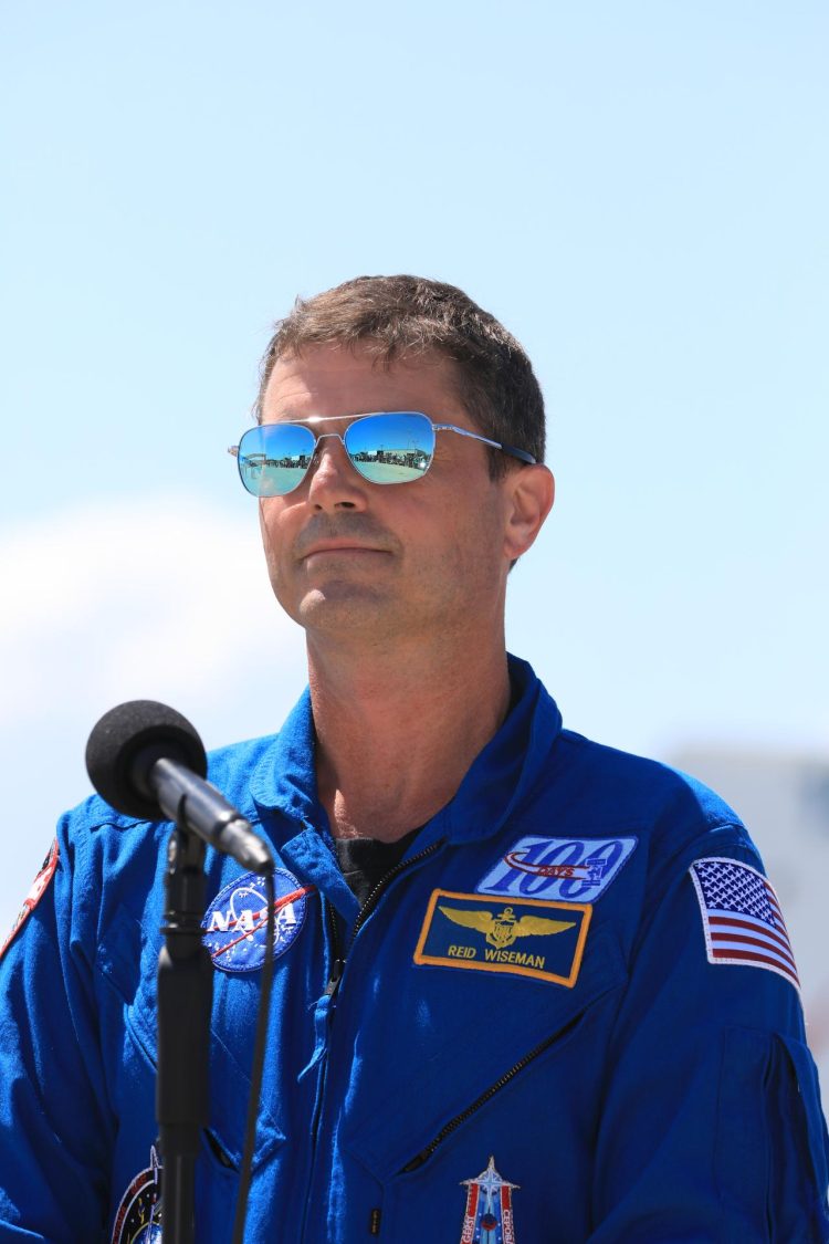 Stojący na tle czystego nieba biały mężczyzna w okularach przeciwsłonecznych i niebieskim mundurze z logo NASA i innymi naszywkami. Przed nim przymocowany na statywie mikrofon.