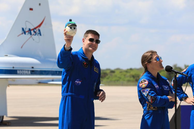 Na pierwszym planie biały mężczyzna w okularach przeciwsłonecznych oraz niebieskim kombinezonie NASA z naszywkami, z uniesioną ręką w której trzyma maskotkę w kształcie białej kuli z zielono-niebieską czapką. Po prawej stronie od niego biała kobieta w okularach przeciwsłonecznych oraz niebieskim kombinezonie NASA z licznymi naszywkami, mówiąca do stojącego przed nią mikrofonu. W tle widać czyste niebo oraz ogon samolotu z logiem NASA.