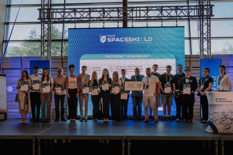 Kilkanaście osób stojących w rzędzie na scenie przed banerem Spaceshield Hack