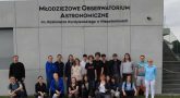 Uczestnicy WWN stojący na trawie pod budynkiem z napisem Młodzieżowe Obserwatorium Astronomiczne im. Kazimierza Kardylewskiego w Niepołomicach.