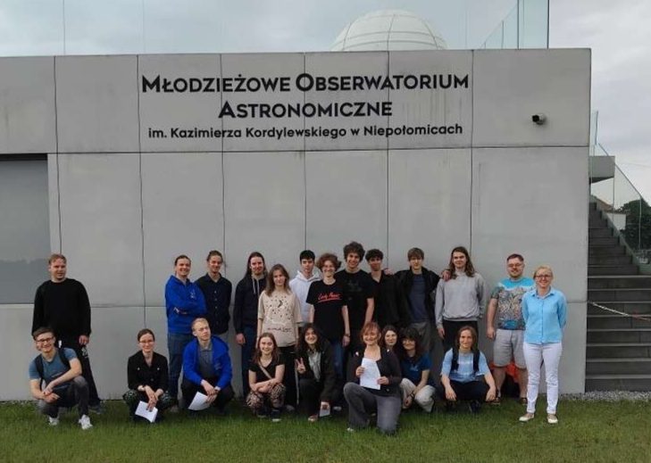 Uczestnicy WWN stojący na trawie pod budynkiem z napisem Młodzieżowe Obserwatorium Astronomiczne im. Kazimierza Kardylewskiego w Niepołomicach.