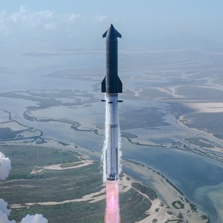 Zdjęcie z lotu ptaka przedstawia rakietę Starship w trakcie lotu. Górny stopień (Ship) o ciemnej, błyszczącej powierzchni ze skrzydełkami aerodynamicznymi jest połączony z jasnym, dolnym stopniem (Super Heavy Booster). Z silników u podstawy rakiety wydobywa się jasnoróżowo-pomarańczowy płomień i kłęby białego dymu. W tle widać rozległą linię brzegową z zatokami, piaszczystymi terenami i wodą pod jasnym, lekko zachmurzonym niebem.