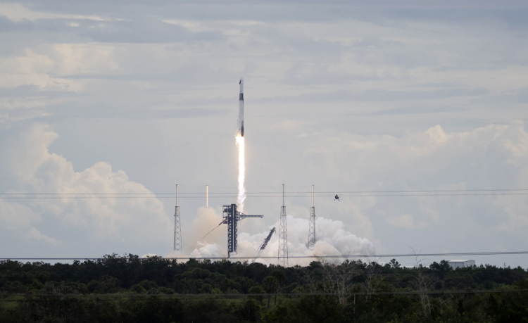 Rakieta SpaceX Falcon 9 unosi się nad wyrzutnią, zostawiając za sobą jasną smugę ognia. U podnóża wieży startowej kłębią się białe obłoki dymu i pary, a w tle widać pochmurne niebo i linię drzew.