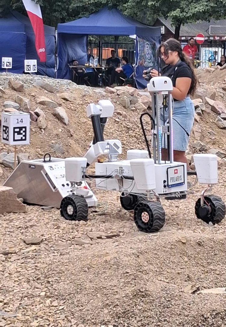 Biały łazik stojący na pomarańczowym, skalistym podłożu, wykonujący czynności ramieniem robotycznym.