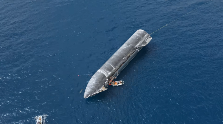 Zdjęcie z lotu ptaka przedstawia stalowo-czarny drugi stopień rakiety Starship dryfujący poziomo w ciemnoniebieskiej wodzie oceanu po próbnym wodowaniu. Widoczne są na nim ślady po przejściu przez atmosferę oraz czarne płytki osłony termicznej. Tuż obok dziobu statku znajduje się mała, biała łódź motorowa z załogą, a w lewym dolnym rogu kadru widać drugą łódź podpływającą do miejsca zdarzenia. Od rufy statku rozciąga się w wodzie lina holownicza.